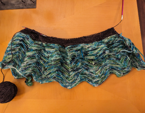A top down photo of a partially knitted blanket. The first finished stripe is a mixture of different blues, the second unfinished stripe is a dark blue