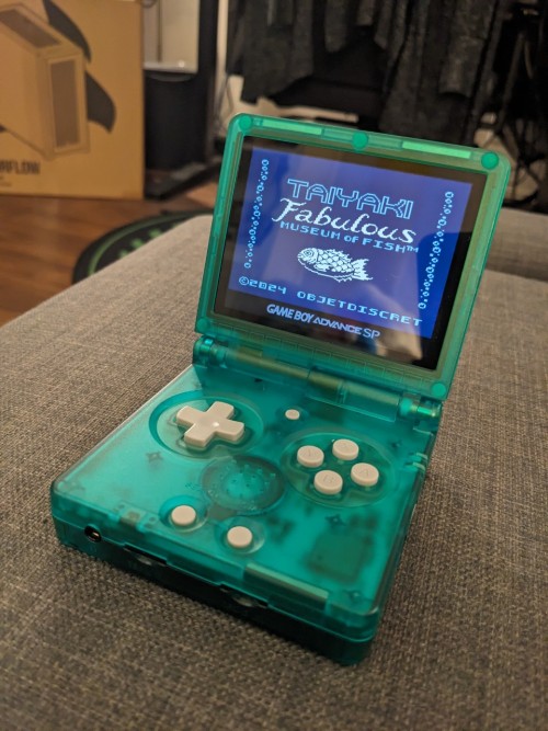 An image of a handheld gaming console that resembles a Game Boy Advance. On it's screen is the titlescreen for the video game, Taiyaki Fabulous Museum of Fish.
